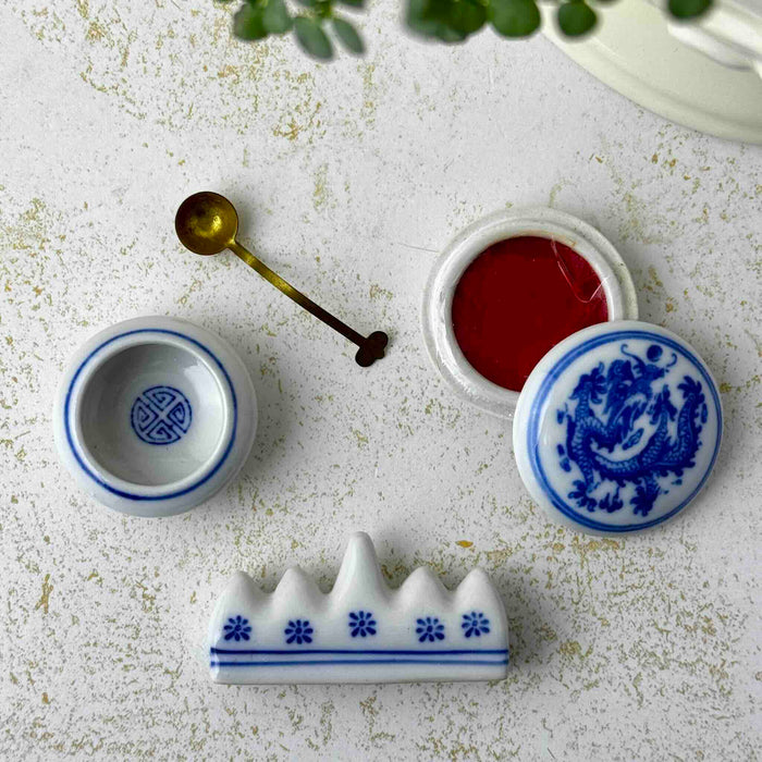 Close-up of vintage Chinese calligraphy water dropper, brass spoon, open porcelain seal paste container, and brush rest on a light surface