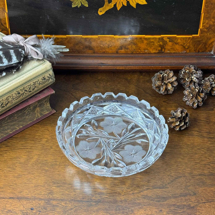 Vintage clear glass floral dish displayed on a dark wooden surface beside stacked books and pinecones, with a decorative wooden panel behind it.