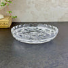 Angled view of the shallow clear glass dish showing its floral center, geometric border, and thick notched rim.