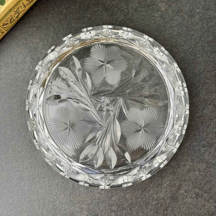 Underside of the round clear glass dish showing the three-flower design, pointed leaves, geometric border, and notched outer edge.
