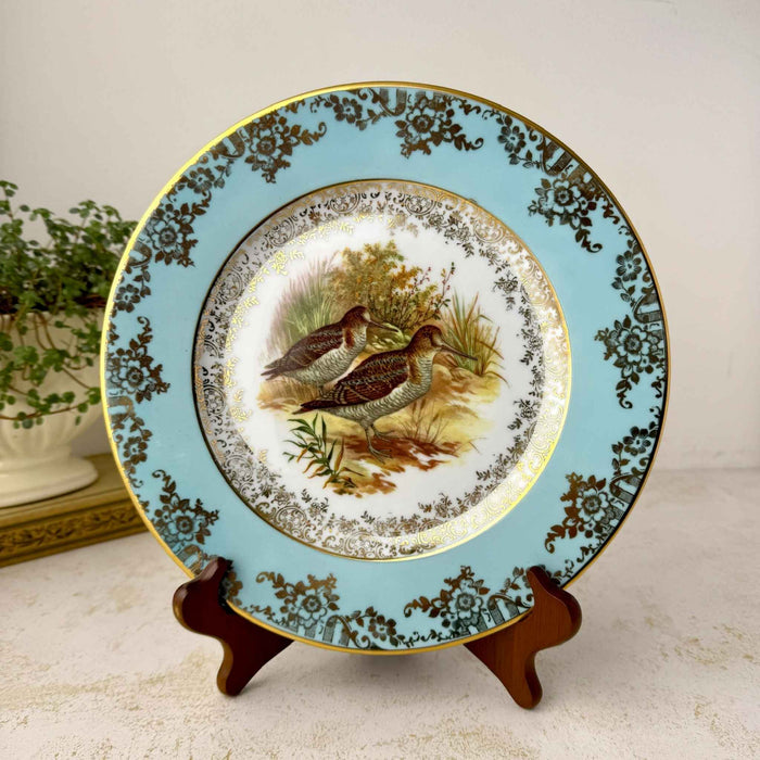 Full front view of a round turquoise and gold game bird plate on a wooden display stand beside a green plant.