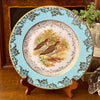 Styled view of a turquoise and gold snipe plate displayed with warm wood, an old book, a trailing plant and a feather.