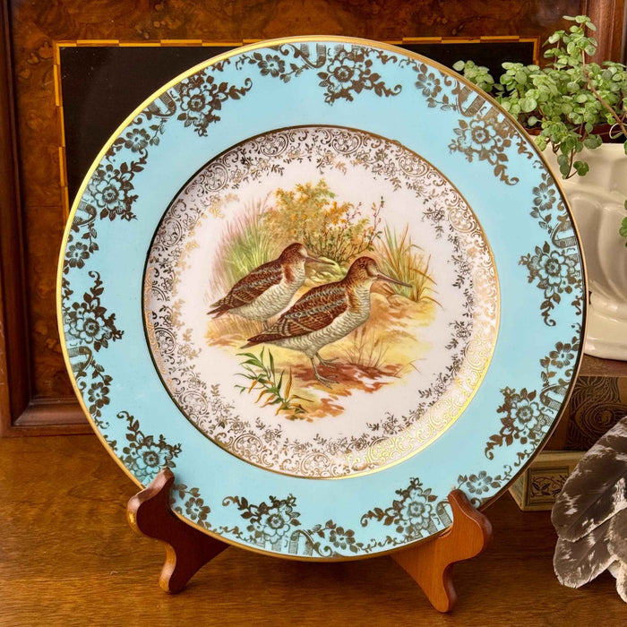 Styled view of a turquoise and gold snipe plate displayed with warm wood, an old book, a trailing plant and a feather.