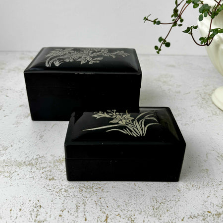 Pair of vintage black lacquer-style decorative boxes with silver botanical motifs and softly domed lids displayed closed on a light textured surface