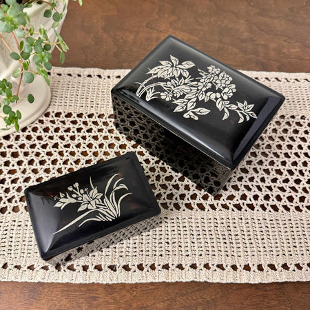 Pair of vintage black lacquer-style trinket boxes with silver floral designs, styled on a crochet runner with trailing plant
