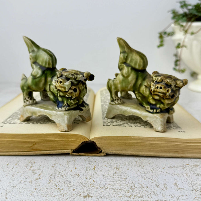 Small vintage green glazed foo dog figurines posed on an open antique book with trailing greenery in the background