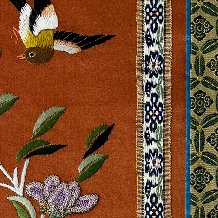 Close-up of vintage Chinese embroidery showing bird, floral motif, patterned trim, and textured stitching on rust-colored fabric panel