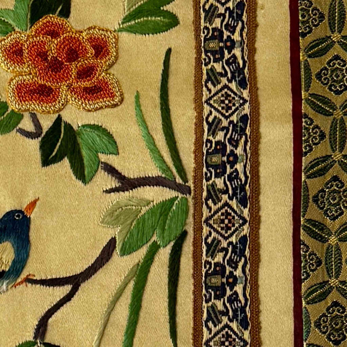 Close-up of vintage Chinese embroidery showing floral branch, bird, patterned trim, and textured stitching on pale gold fabric panel
