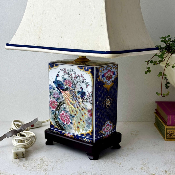 Three-quarter view of a vintage ceramic peacock accent lamp with original shade, shown beside stacked antique books against a neutral wall