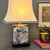 Vintage chinoiserie peacock lamp styled on a table with brass planter, artwork, and pink book