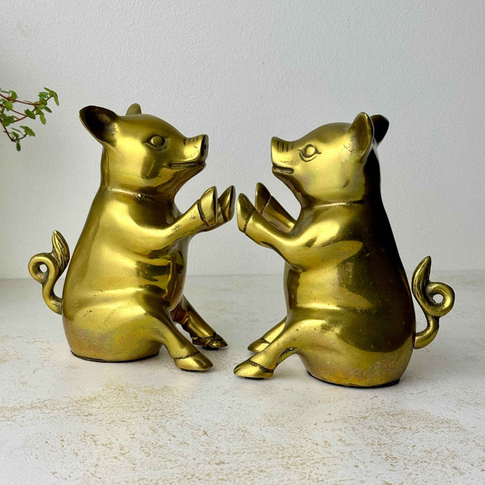 Pair of vintage brass pig bookends facing each other, showing their seated forms, raised hooves, pointed ears, and curled tails.