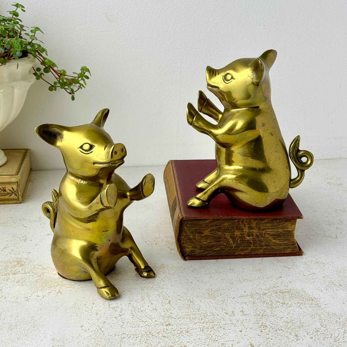 Pair of vintage brass pig bookends seated with raised front hooves and curled tails, one displayed on an old book.