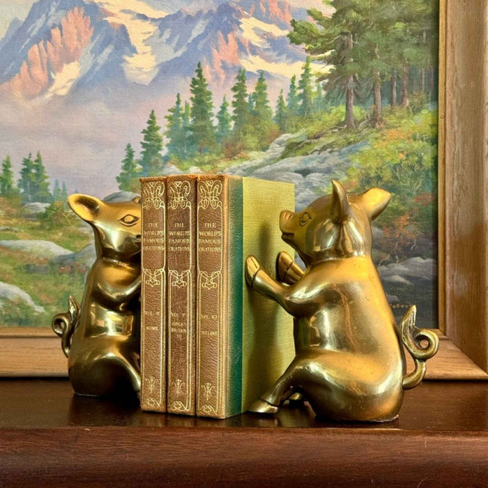 Vintage brass pig bookends holding three small books on a wooden shelf in front of framed landscape artwork.