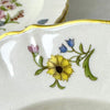 Close-up of a small floral spray with a yellow flower, blue blossoms, pink bud, and green leaves beside the gold-trimmed rim.