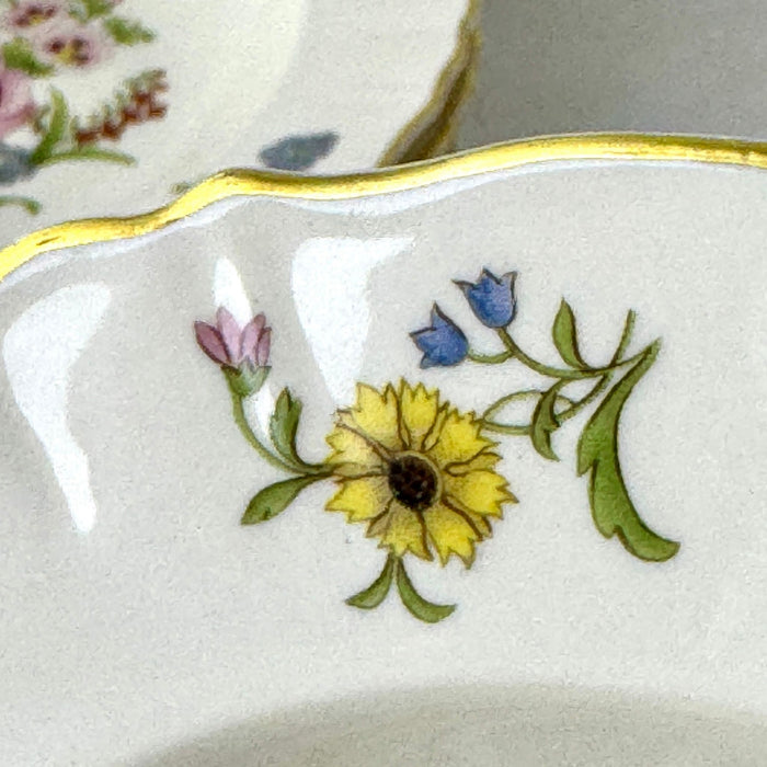 Close-up of a small floral spray with a yellow flower, blue blossoms, pink bud, and green leaves beside the gold-trimmed rim.
