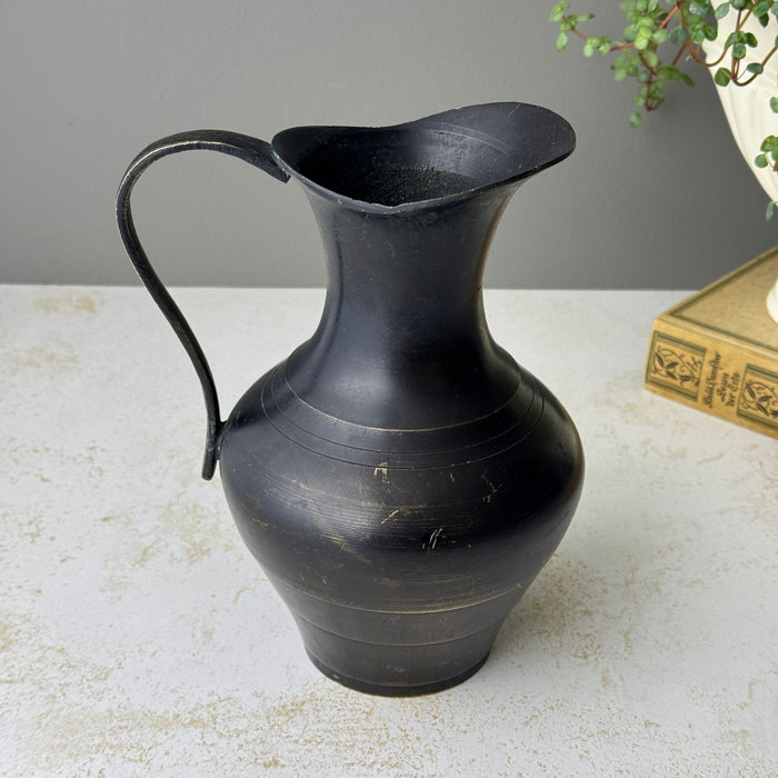 Opposite side of vintage blackened brass pitcher showing rounded body, flared lip, curved handle, and surface wear.
