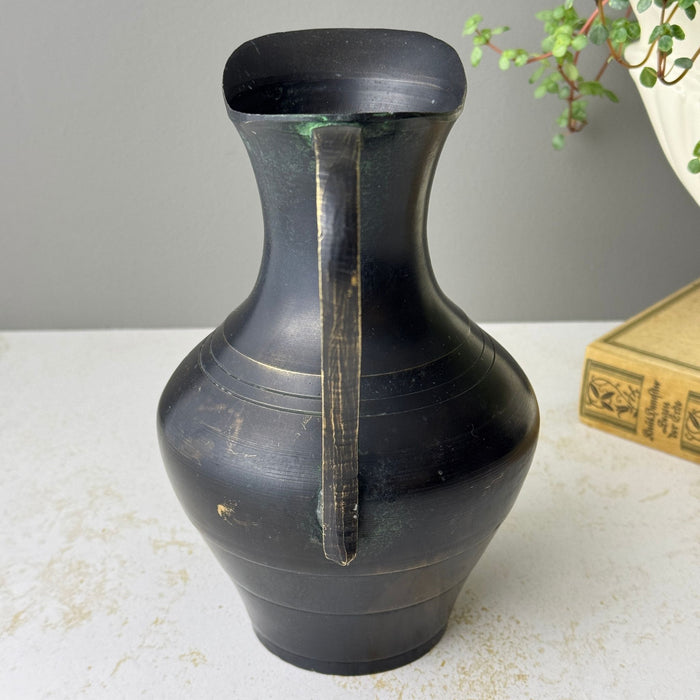 Rear view of blackened brass pitcher showing the long narrow handle attached to the neck and rounded body.