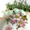 Close-up of mauve floral decoration with raised gold accents, green leaves, aqua shading, and gilded scalloped edge.