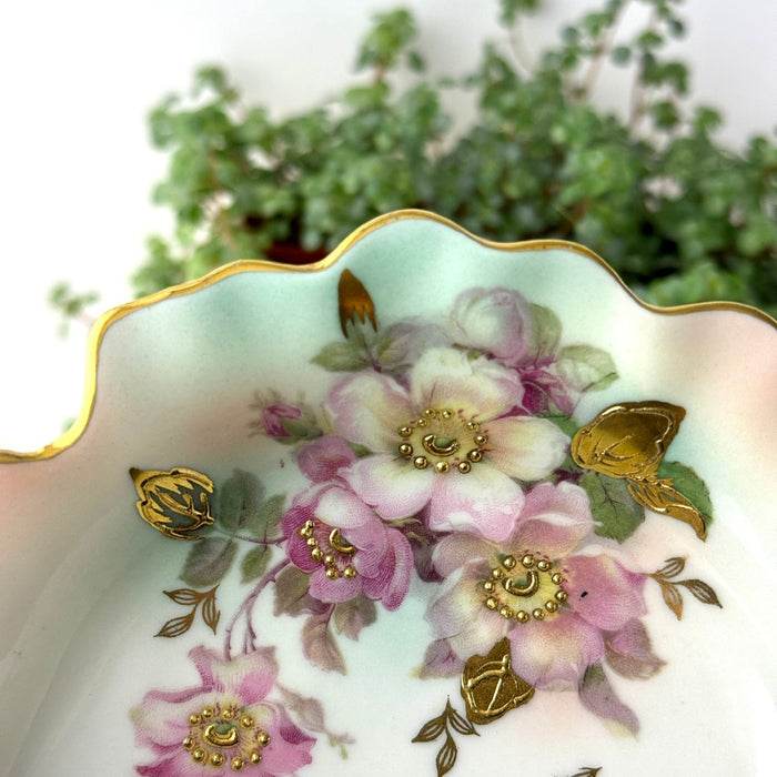 Close-up of mauve floral decoration with raised gold accents, green leaves, aqua shading, and gilded scalloped edge.