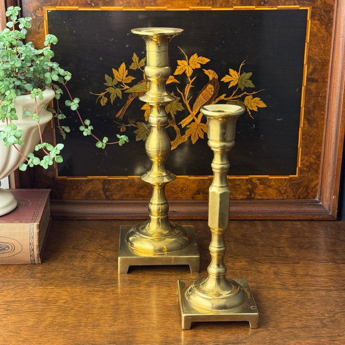 Two vintage brass candlesticks with staggered heights styled on a wood surface in front of a decorative inlaid bird and foliage tray.