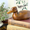 Vintage carved wood duck styled on stacked old books beside a plant and bird artwork.