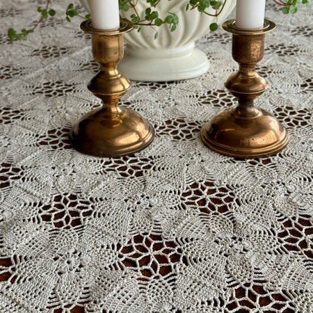 Close view of flax beige crochet tablecloth spread over dark wood, with brass candlesticks above the repeating floral rosette and openwork pattern.