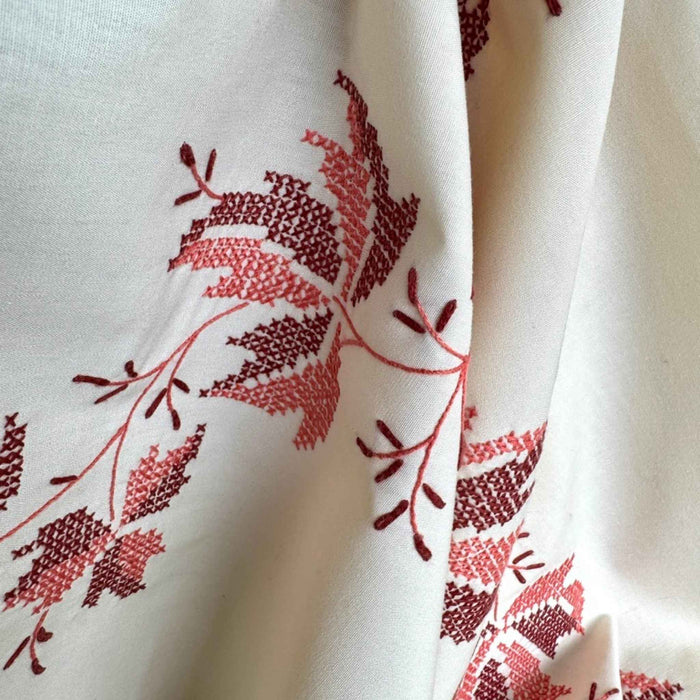 Close-up of the warm cream tablecloth falling in soft folds, showing its fluid drape and burgundy and coral embroidered autumn leaves.