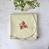 Overhead view of the folded cream tablecloth with an embroidered burgundy and coral leaf motif and taupe scalloped edge.
