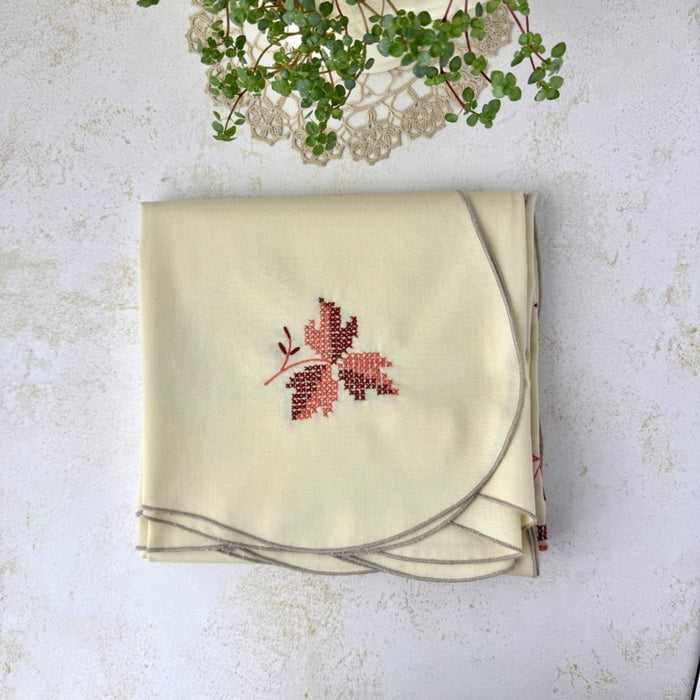 Overhead view of the folded cream tablecloth with an embroidered burgundy and coral leaf motif and taupe scalloped edge.