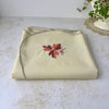 Folded vintage tablecloth viewed from a low angle, showing the cream fabric, embroidered leaf motif and softly scalloped edging.