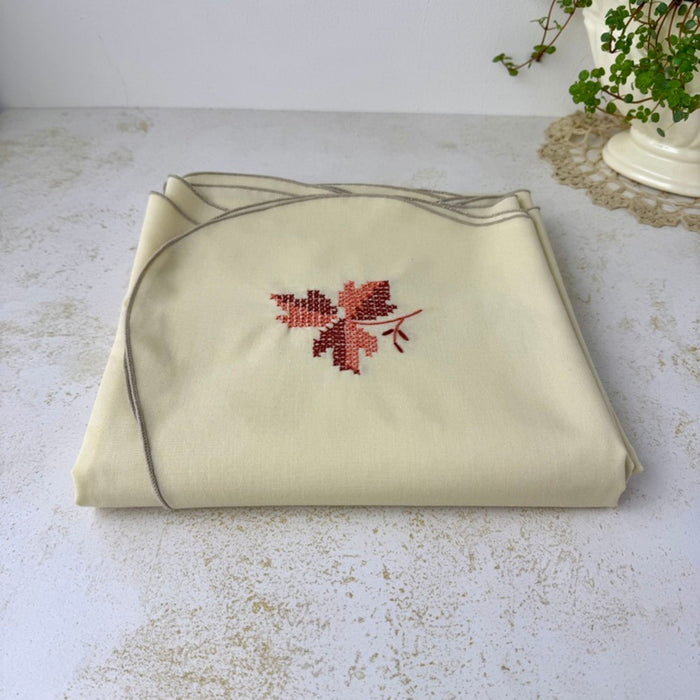Folded vintage tablecloth viewed from a low angle, showing the cream fabric, embroidered leaf motif and softly scalloped edging.
