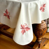 Warm cream embroidered tablecloth draped over a wooden table, showing scattered burgundy and coral leaf motifs and rounded corners.
