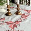 Vintage embroidered tablecloth spread across a dining table with burgundy and coral autumn leaves, brass candlesticks and white candles.