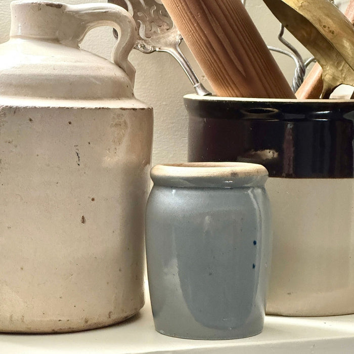 Small gray-blue stoneware crock displayed on a kitchen shelf between larger vintage utilitarian pottery vessels.