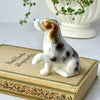 Small hand-painted ceramic dog figurine with a raised front paw, brown and gray markings, displayed on a vintage book.