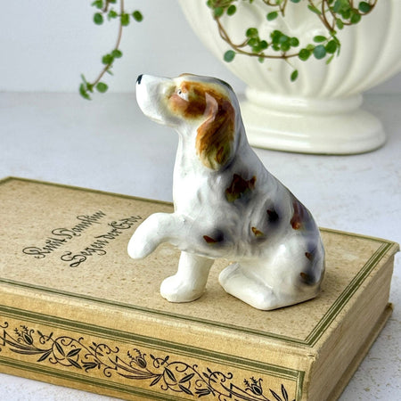 Small hand-painted ceramic dog figurine with a raised front paw, brown and gray markings, displayed on a vintage book.