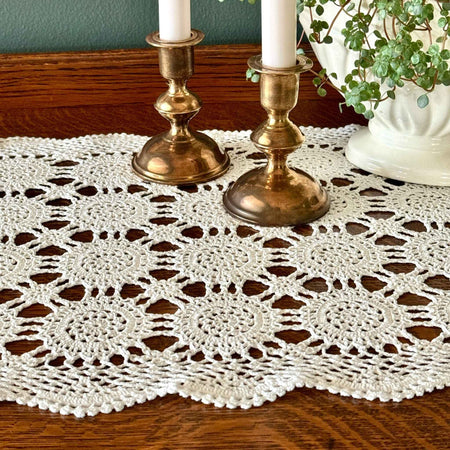 Ecru crochet table runner styled across a dark wood table with brass candlesticks, white candles, and a white planter with greenery.