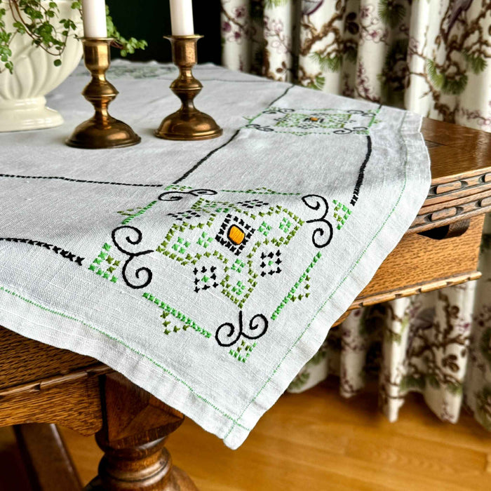 Ecru square embroidered tablecloth draped over a dark wood table, showing navy and green geometric medallions with small orange centers, styled with brass candlesticks and a white planter.