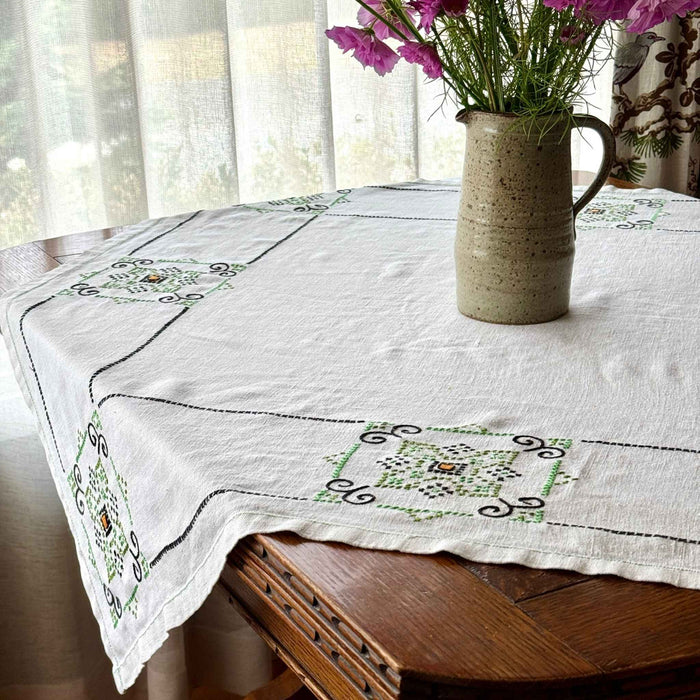 Square ecru embroidered tablecloth spread over a dark wood dining table, showing repeating navy and green geometric designs and an orange-centered motif, styled with a pottery pitcher of flowers.