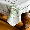 Close view of one corner of the ecru tablecloth draped over a dark wood table, showing the green, navy, and orange cross-stitch and embroidered design.