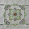 Close-up of a geometric embroidered medallion worked in two shades of green, navy, and orange on the ecru woven cloth.