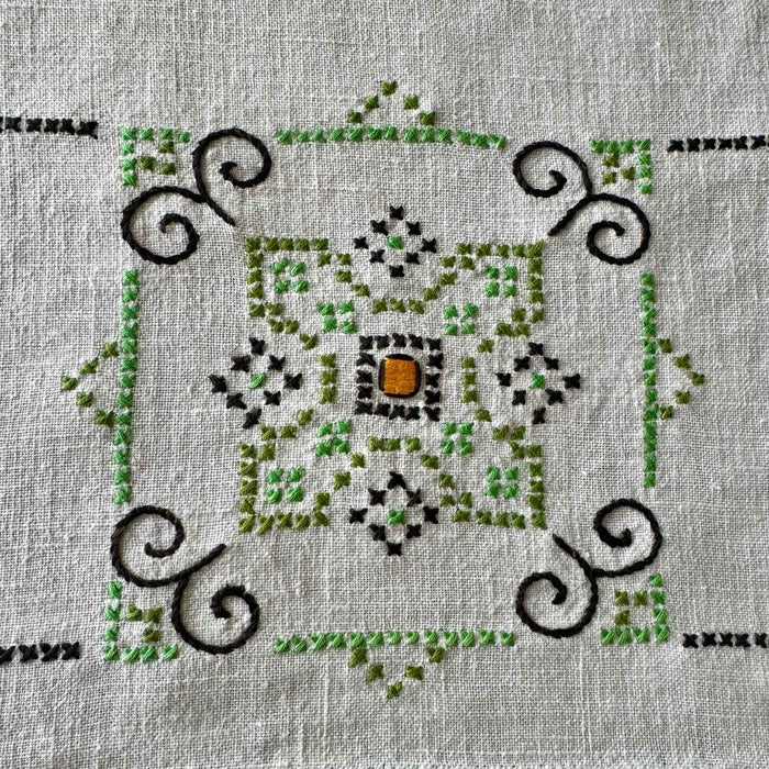 Close-up of a geometric embroidered medallion worked in two shades of green, navy, and orange on the ecru woven cloth.