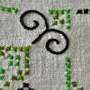 Detail of the embroidery showing navy scrolling lines and green cross stitches on the textured ecru woven fabric.