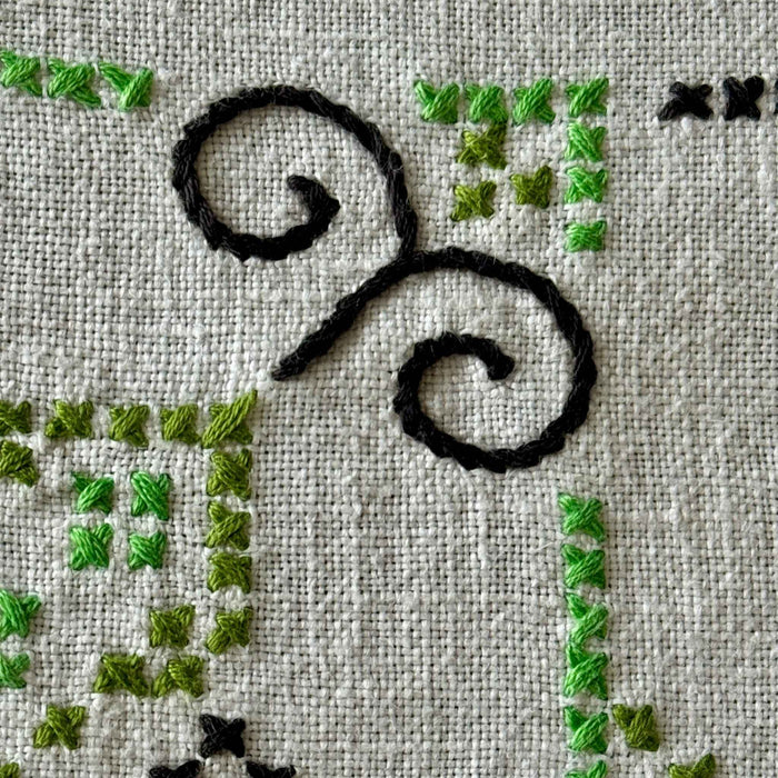 Detail of the embroidery showing navy scrolling lines and green cross stitches on the textured ecru woven fabric.
