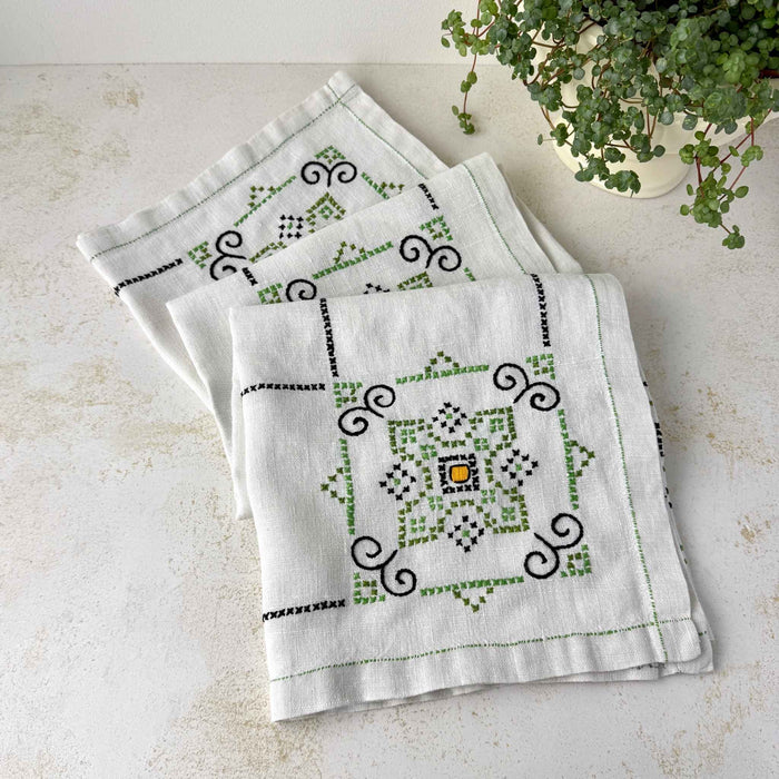 Ecru embroidered tablecloth loosely folded on a light surface, showing several repeating green, navy, and orange geometric medallions beside a potted plant.