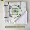 Folded ecru embroidered tablecloth with a corner turned back over a white linen for color comparison, showing a green, navy, and orange medallion.
