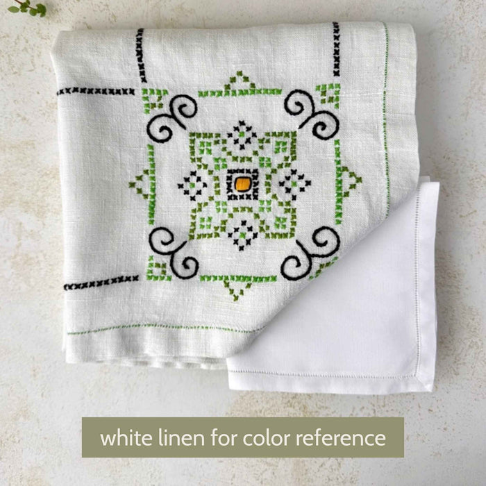 Folded ecru embroidered tablecloth with a corner turned back over a white linen for color comparison, showing a green, navy, and orange medallion.