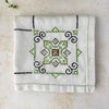 Folded ecru embroidered tablecloth on a light surface, showing one geometric medallion in green, navy, and orange.