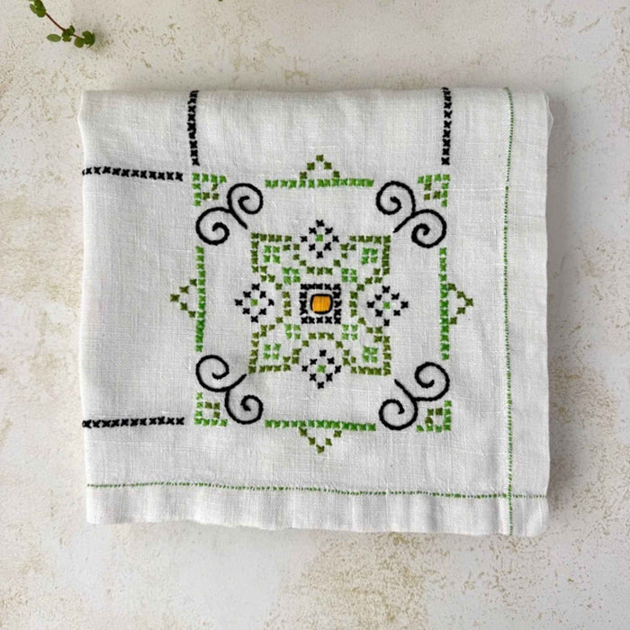 Folded ecru embroidered tablecloth on a light surface, showing one geometric medallion in green, navy, and orange.