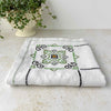 Low angle folded view of ecru embroidered tablecloth on a light surface, showing one geometric medallion in green, navy, and orange beside a potted plant.